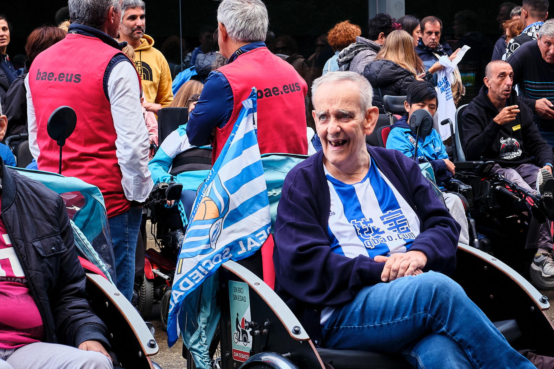 Un hombre sonriente en un triciclo con personas en ellos, con indumentaria de la Real Sociedad, rodeados de viandantes, frente al campo de Anoeta