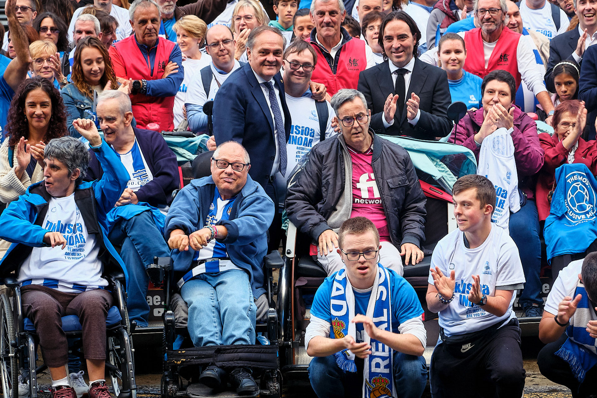 Varios triciclos con personas en ellos, con indumentaria de la Real Sociedad, rodeados de viandantes, frente al campo de Anoeta